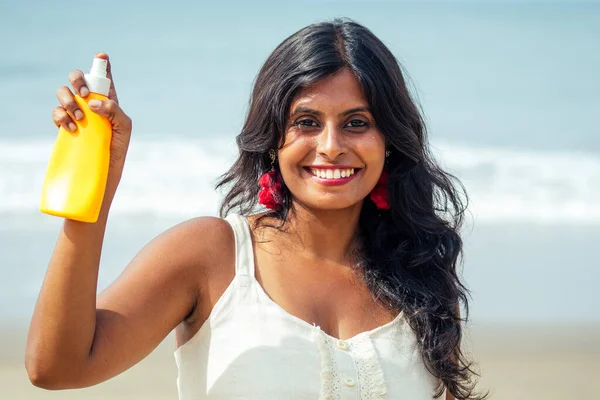 Retrato de una hermosa y sonriente sonrisa blanca como la nieve mujer india pelo rizado negro y piel oscura en una camiseta blanca sosteniendo botella de protector solar aerosol en beach.girl disfrutando de vacaciones spf cuerpo paraíso —  Fotos de Stock