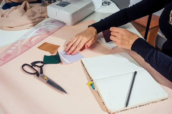 Bela costureira com cabelo longo escreve em um caderno. o alfaiate cria uma coleção de roupas. jovem mulher roupas de grife trabalhando com couro — Fotografia de Stock