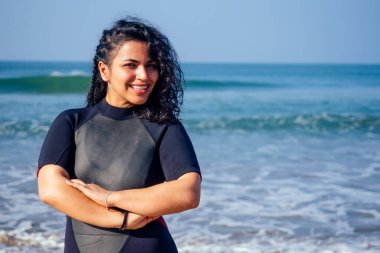 Siyah dalgıç kıyafeti giymiş Hintli sörfçü elleri çapraz suya yakın duruyor.