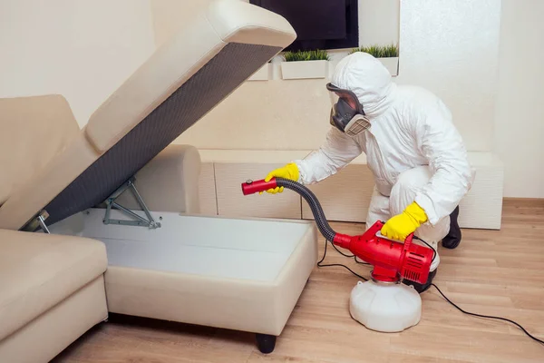 Trabalhador de controle de pragas em pesticidas de pulverização uniforme sob o sofá na sala de estar — Fotografia de Stock