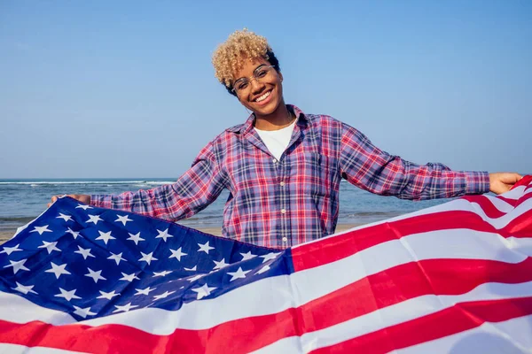 Heureuse femme afro américaine tenant le drapeau des Etats-Unis à la plage tropicale ensoleillée.4 juillet Independence Day concept — Photo