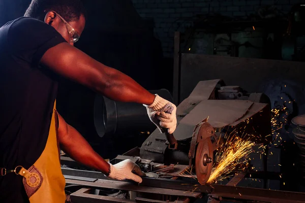 Een knappe Afrikaans-Amerikaanse man die staal smeed naast een oven in een donkere werkplaats. kleine ondernemingen, — Stockfoto