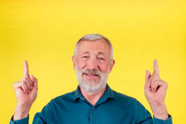 Retrato de primer plano de un anciano feliz mirando a la cámara, usando una camisa de algodón verde con un cuello apuntando con el dedo hacia arriba para copiar el espacio en el fondo amarillo del estudio —  Fotos de Stock