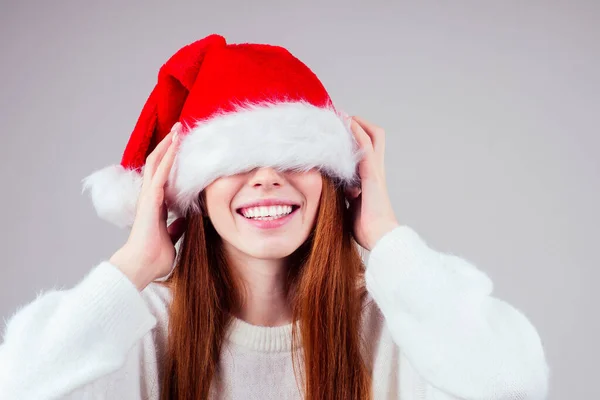 Blyg vacker rödhårig ingefära kvinna bär vit stickad tröja och gömmer ögat i Santa claus hatt studio bakgrund — Stockfoto