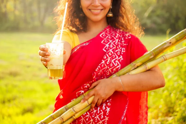 Красивая индуистка в красном сари, стоящая рядом с аппаратом сахарного тростника производителя сока, плантации летнего фермерского фона. создание малого бизнеса — стоковое фото