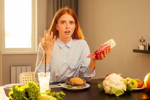 红头发的姜汁女孩在家里的厨房里吃着带有面包芝麻和塑料薄膜垃圾的假汉堡包，喝着装有非生态管的一次性杯子里的水 — 图库照片