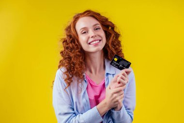 Kızıl saçlı, kıvırcık saçlı, beyaz iş kadını pamuklu gömlek giyiyor, kredi kartı stüdyosu sarı arka plan kara Cuma indirimi yapıyor.