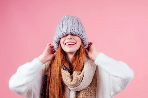 Tímida ruiva mulher gengibre colocando um chapéu de malha em sua cabeça, escondendo olho e neve branco sorriso boca no estúdio rosa fundo — Fotografia de Stock
