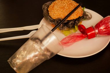 Bun susamlı sahte burger ve plastik selofan film çöpü. Ekolojik olmayan bir bardaktan içme suyu. Ev mutfağının kapkaranlık arka planında çatal bıçak.