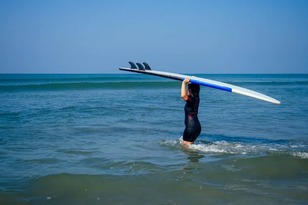Indianerin im Taucheranzug verbringt Zeit am Meer von Goa. Sie trägt Surfbrett auf dem Kopf — Stockfoto