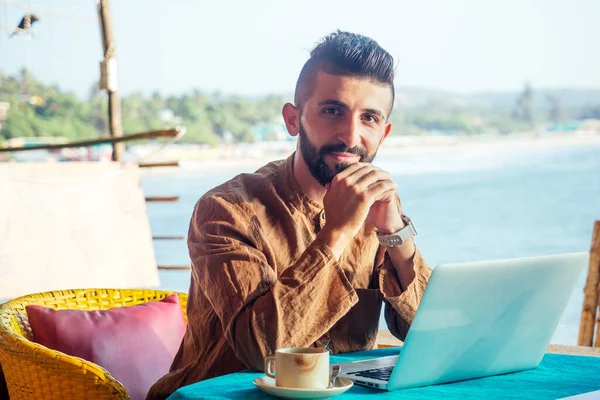 Attractive egyptian male worker is using computer in cafeteria with sea background — Stock Photo, Image