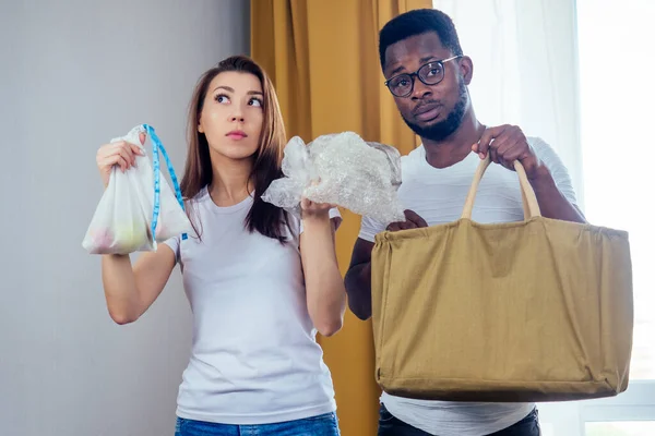 Coppia multinazionale che sceglie tessile o poliestere bag.african americano uomo e donna asiatica salvare pianeta in piedi in salotto — Foto Stock