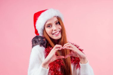 Mutlu kızıl saçlı kadın Noel Baba şapkası takıyor ve sevimli siyah fareye sarılıyor ve el stüdyosunun pembe arka planından kalp şekli yaratıyor.
