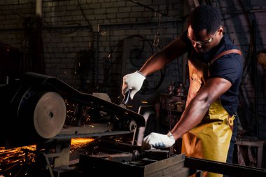 handsome african americam man forging steel next to furnace in dark workshop. small business comcept clipart