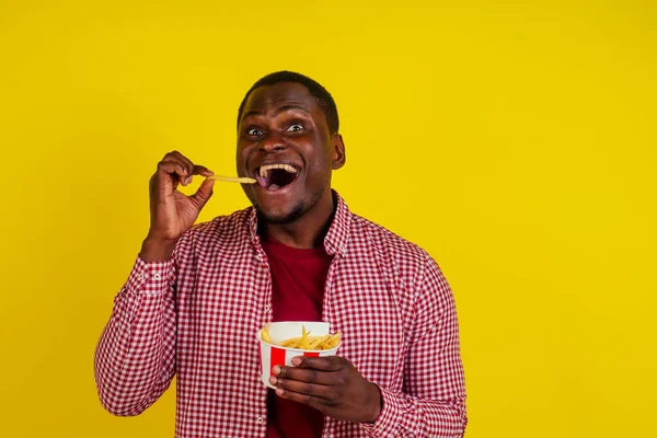 Pohledný latin muž jíst hranolky s chutí k jídlu a požitek ve studiu žluté pozadí — Stock fotografie