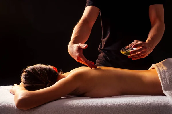 Молодая индийская женщина лежит на столе и получает аюрведический массаж с органическим маслом или медовый в темной room.massagist мужчина наливая клиента обратно — стоковое фото