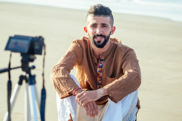 Арабский мужчина сидит на песке и говорит штатив видео решений на пляже. Концепция создания контента для путешествий — стоковое фото