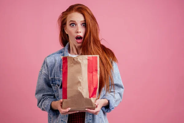 Нахмурившись удивлен и изумлен длинные рыжие волосы рыжая женщина держит бумажный пакет эко-мешок с чипсами в студии розовый background.she не хочет есть нездоровую пищу — стоковое фото