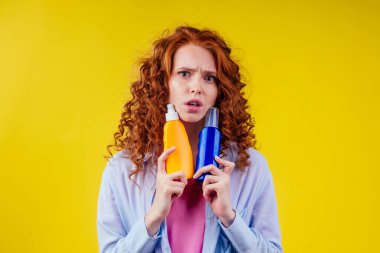 Kızıl saçlı kıvırcık saçlı kadın elinde güneş kremi şişesiyle stüdyo sarı arka planda doğru seçimi yapıyor.