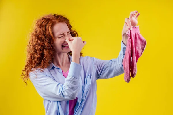 Pelirroja pelirroja rizada con un gesto de camisa de algodón huele mal sosteniendo un calcetín rosa sucio hacia fuera una mirada asquerosa en su cara en fondo amarillo estudio — Foto de Stock