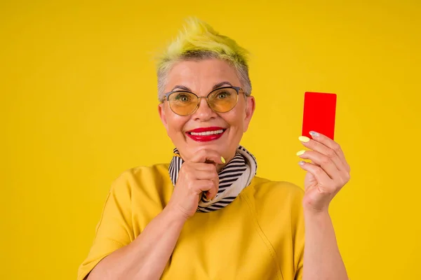 Mogen gammal kvinna med färgade snygga gula färgat hår och röda läppstift läppar i varm klänning med kreditkort bakgrund studio: svart fredag försäljning — Stockfoto