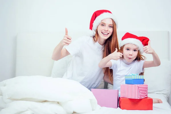 Vacker rödhårig ingefära kvinna med tomte klo hatt och glad liten flicka i pyjamas sitter på sängen visar tummen upp gest bredvid gåvor boxes.christmas morgon firande — Stockfoto