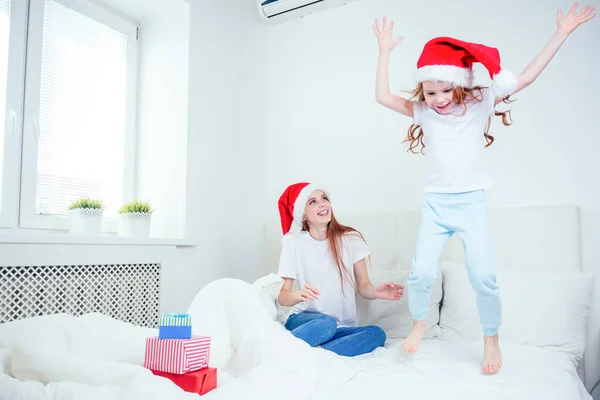 Vacker rödhårig ingefära kvinna med Santa claus hatt och glad liten flicka i pyjamas hoppar på sängen med vit kudde och filt bredvid gåvor boxes.christmas morgon firande — Stockfoto