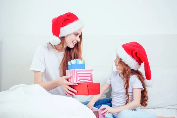 Vacker rödhårig ingefära kvinna med Santa claus hatt och glad liten flicka i pyjamas sitter på sängen med vit kudde och filt bredvid gåvor boxes.christmas morgon firande — Stockfoto