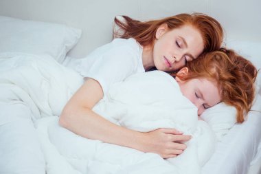 Güzel kızıl saçlı kadın, sabah evinde beyaz battaniyenin altında uyuyan kızını kucaklıyor. Corona enfeksiyonu salgını.