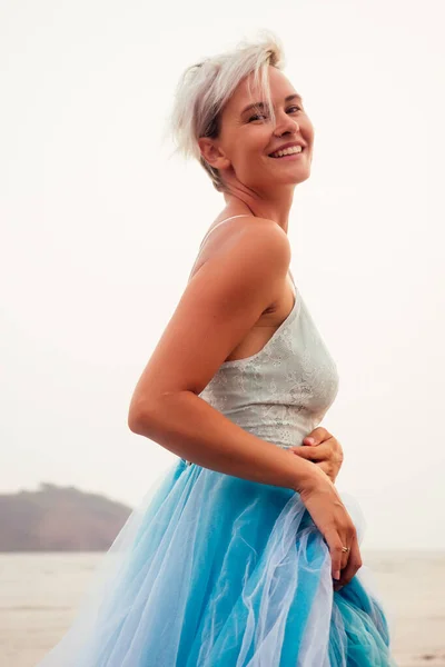 Hermosa rubia mujer de pelo corto posando en tul azul vestido de tren largo en la playa de arena negra — Foto de Stock