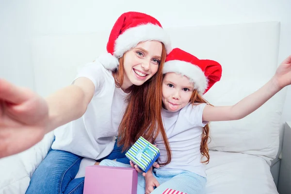 Vacker rödhårig ingefära kvinna i Santa claus hatt och glad liten flicka i pyjamas sitter på sängen med presentaskar, ta selfie bilder på smartphone camera.christmas morgon firande — Stockfoto