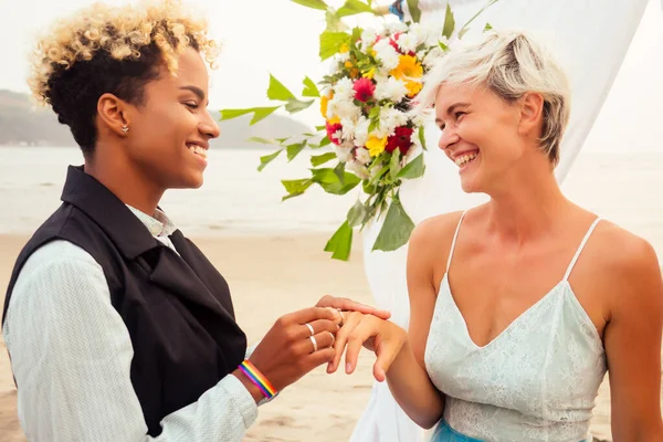 Noivo feminino afro-americano em terno preto e noiva loira caucasiana feliz amando juntos na cerimônia de praia sob arco floral — Fotografia de Stock