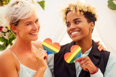 Siyah takım elbiseli Afro-Amerikan kadın damat ve düğün çiçekli kemerin altındaki tropikal plajda resmi giyinmiş beyaz gelin.