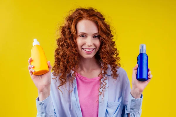 Kızıl saçlı kıvırcık saçlı kadın elinde güneş kremi şişesiyle stüdyo sarı arka planda doğru seçimi yapıyor.. — Stok fotoğraf