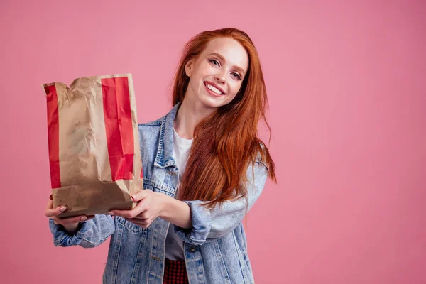 Happy long red-haired ginger woman holding paper eco bag package with chips in studio pink background — Stock Photo, Image