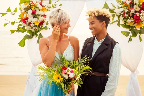 afro american female in the role of the groom and short blonde haired bride in blue dress in ceremony under wedding arch in tropical beach