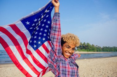 Güneşli tropik plajda bayrak tutan mutlu Afro-Amerikan kadını. 4 Temmuz Bağımsızlık Günü konsepti.