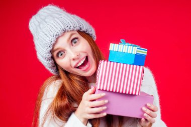 Kızıl saçlı kadın şık yün şapka ve beyaz kazak giyer. Kameraya bakar ve üç kutunun arkasına saklanır. Stüdyo kırmızı arka planda hediyeler.