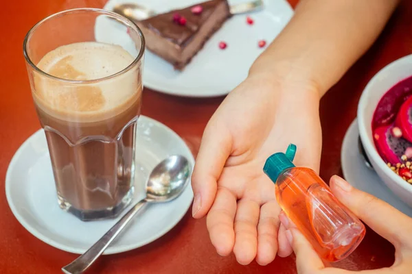 woman useing antibacterial gel on hands before start to eating her healthy food in tropical asian cafe . smoothie bowl , raw cake and coffee on table with sanitizer on bottle