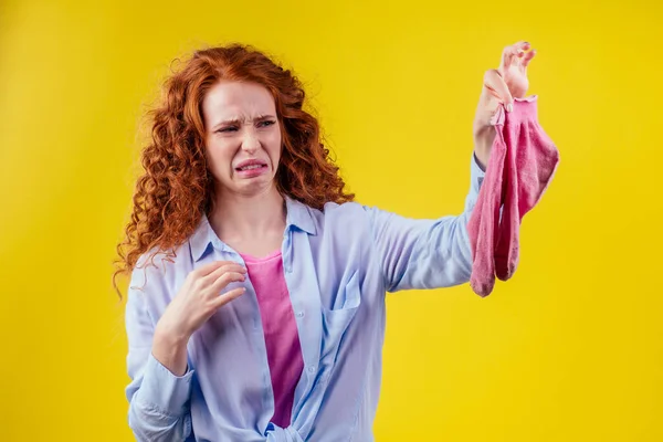 Кудрявая рыжая женщина в хлопчатобумажной рубашке жест плохо пахнет держа грязный розовый носок из отвратительный взгляд на ее лице в студии желтый фон — стоковое фото