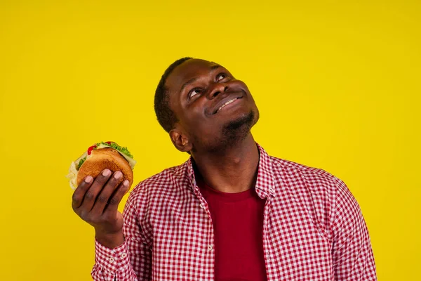 Jovem afro-americano homem comendo hambúrguer isolado no fundo amarelo olhando para cima — Fotografia de Stock