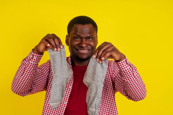 Homem afro-americano insatisfeito fecha o nariz, sente sobre meias cheiro ruim, veste camiseta vermelha casual em estúdio fundo amarelo — Fotografia de Stock