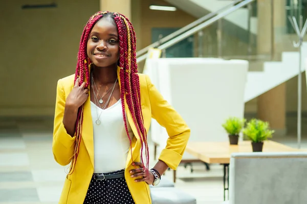 Glückliche, afroamerikanische Frau in stylischem gelben Sakko und bunten Dreadlocks mit Zöpfen am Arbeitsplatz in einem hellen, großen modernen Büro — Stockfoto