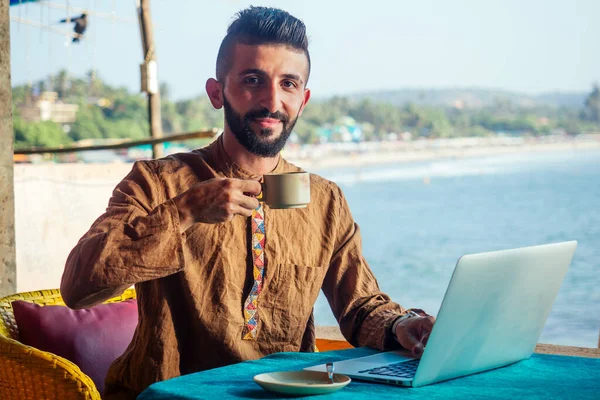 Egyptian happy man with laptop computer drinking coffee or masala chai at beach shack in Goa