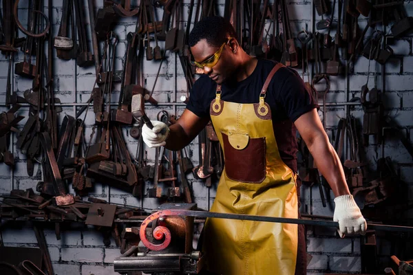 Een knappe Afrikaans-Amerikaanse man die staal smeed naast een oven in een donkere werkplaats. kleine ondernemingen, — Stockfoto