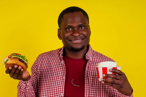 Bonito latina homem comer batatas fritas pepitas e hambúrguer com apetite e prazer no estúdio amarelo background.chicken pernas em um balde — Fotografia de Stock