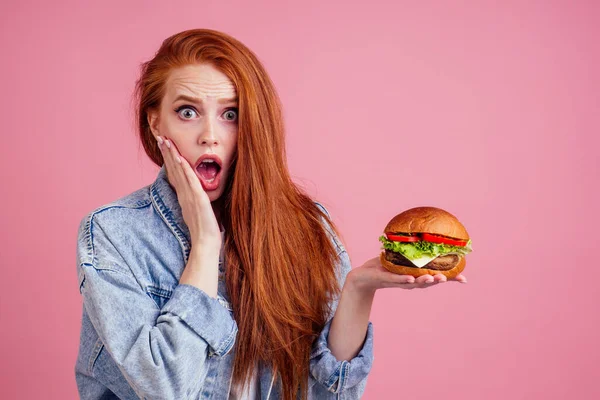 Bella zenzero dai capelli rossi rossa donna in possesso di grande cheeseburger con formaggio, costoletta di manzo e lattuga di pomodoro in studio sfondo rosa .. — Foto Stock