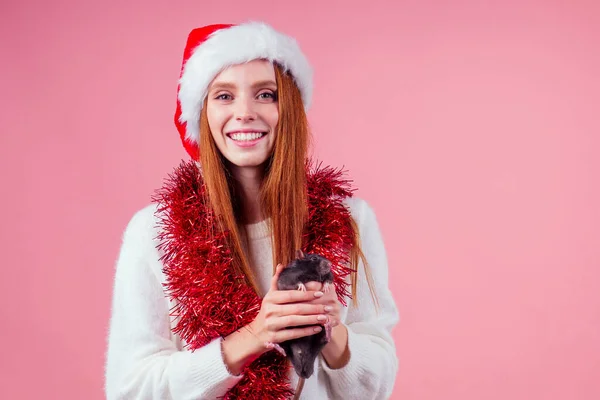 Glad rödhårig ingefära kvinna bär Santa claus hatt och krama söt svart råtta i studio rosa bakgrund — Stockfoto