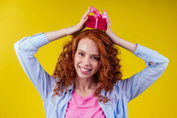 Rothaarige, lockige Ingwerfrau mit Geschenkschachtel auf dem Kopf im Atelier vor gelbem Hintergrund. Muttertag und achter Valentinstag des Marchors — Stockfoto