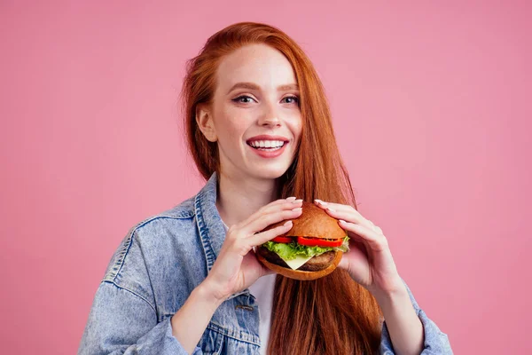 Rothaarige Ingwerfrau mit Sommersprossen genießt große riesige Burger Schnitzel und trägt demin amerikanischen Jeansjacke im Studio rosa Hintergrund .USA traditionelles Konzept — Stockfoto